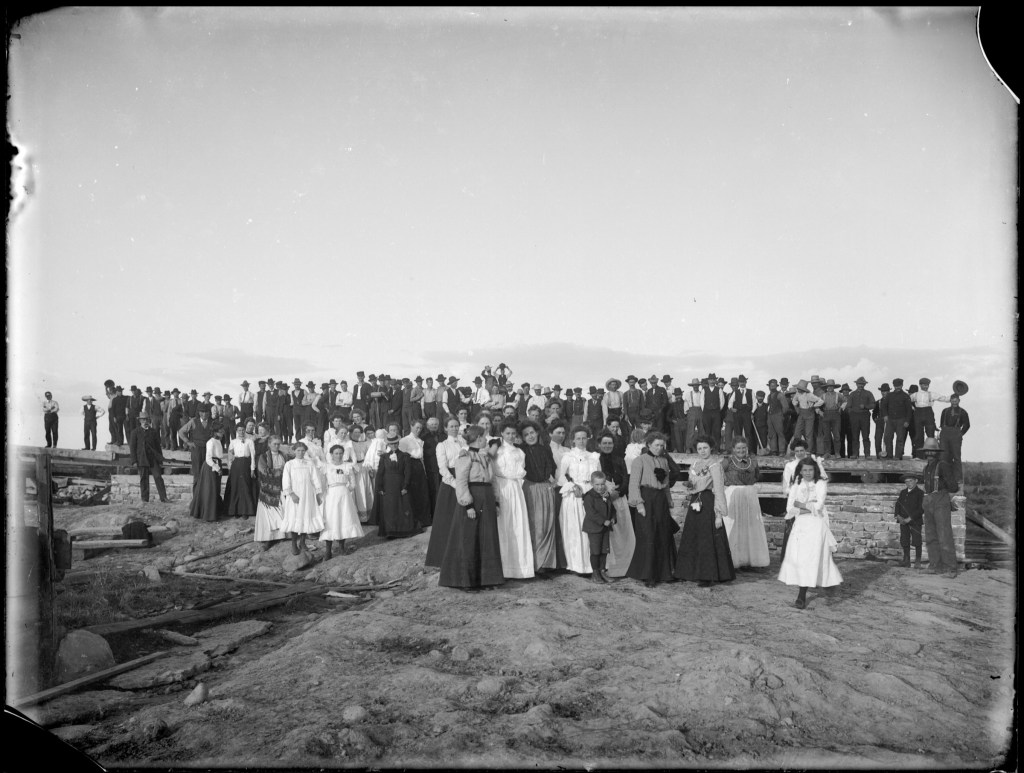 Several dozen neighbours standing on and around the timber frame of a half-raised barn, early 1900s.